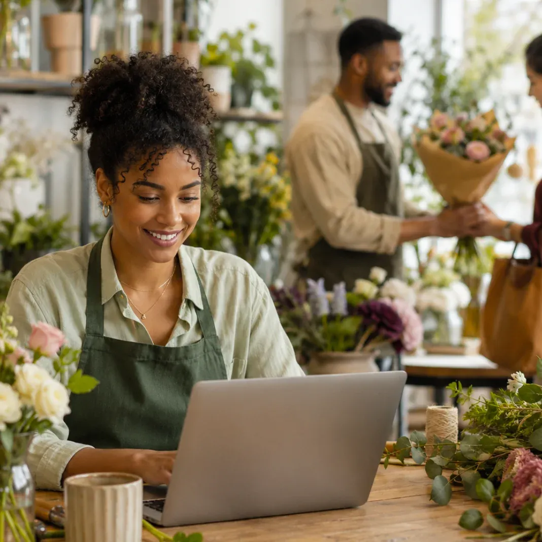 A smiling small business florist managing her shop online on a laptop while a colleague serves a customer