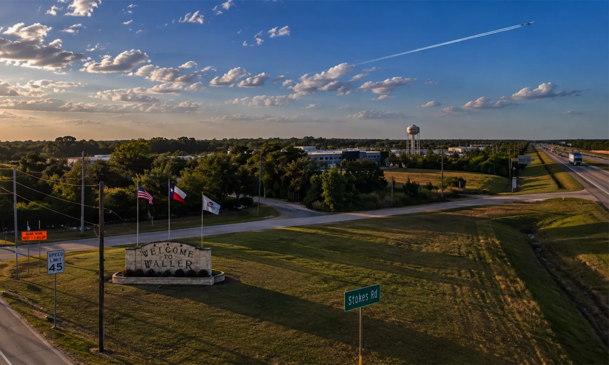 Aerial view of the Welcome to Waller stone sign with U.S. and Texas flags, the town water tower and Highway 290 beyond, at golden hour